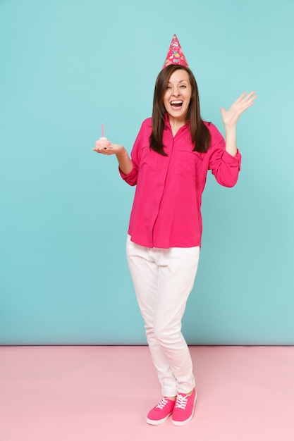 Mulher de retrato de corpo inteiro em blusa de camisa rosa, calça branca, chapéu de aniversário com cupcake posando isolado na parede pastel azul rosa brilhante.