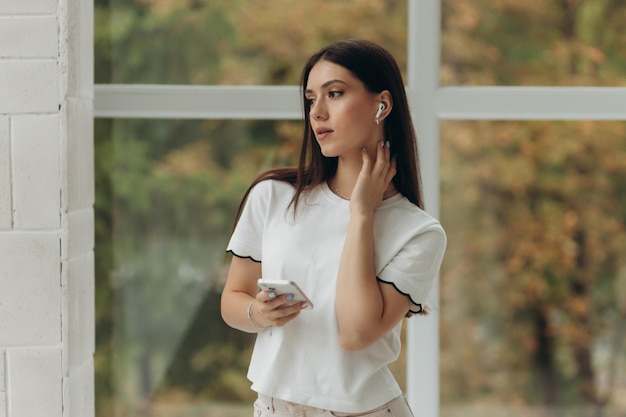 Mulher de retrato fotográfico de camisa branca usando fones de ouvido sem fio curtindo música relaxante em casa