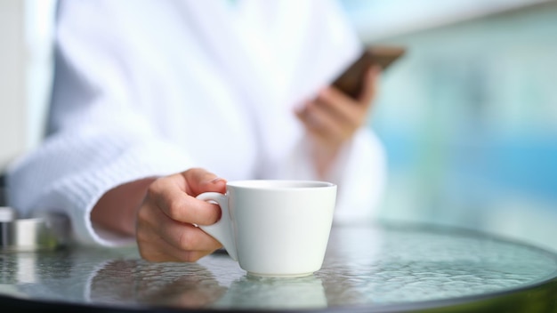 Mulher de roupão com telefone celular nas mãos bebendo café de xícara branca à mesa em spa closeup