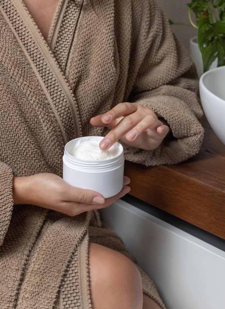 Foto mulher de roupão de banho castanho tocando creme com o dedo e segurando um frasco de cosméticos no banheiro