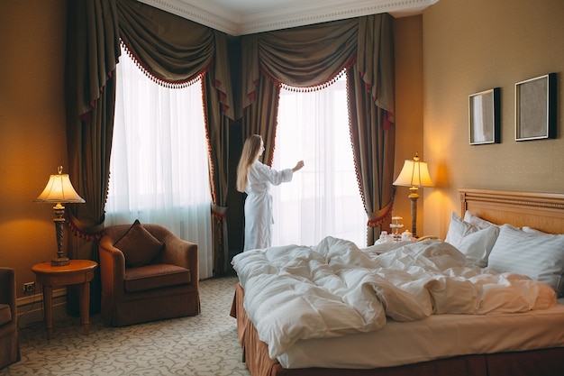 Mulher de roupão fica perto da janela no quarto de hotel.