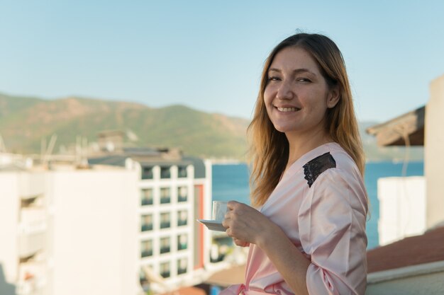 Mulher de roupão tomando café na varanda de seu quarto no hotel