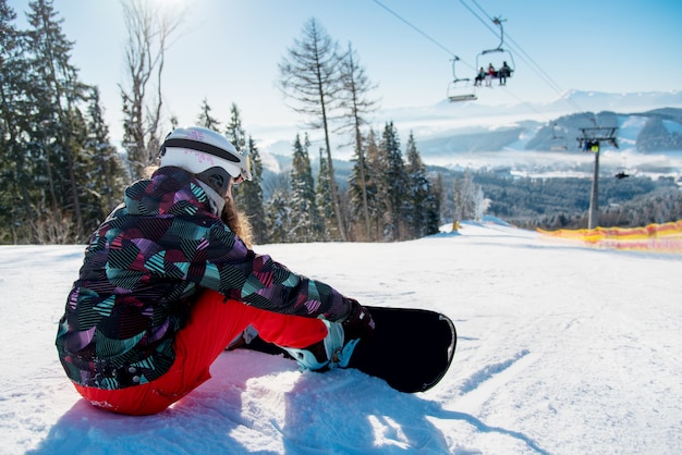 Mulher de snowboarder descansando na pista de esqui sob o teleférico com uma bela paisagem das montanhas e florestas em um dia ensolarado