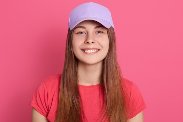 Mulher de sorriso feliz que olha brincalhão ao levantar isolado sobre a parede cor-de-rosa, vestindo a camisa vermelha de t e o boné de beisebol.