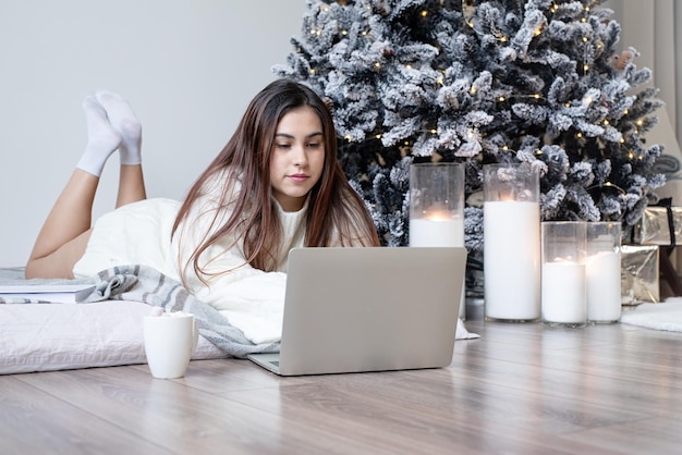 Mulher de suéter branco quente de inverno deitada na cama em casa usando laptop