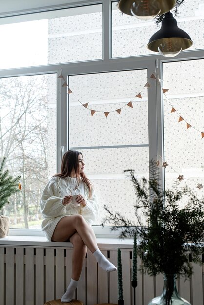 Mulher de suéter branco quente de inverno sentada no parapeito da janela em casa na véspera de Natal segurando uma xícara com marshmallows