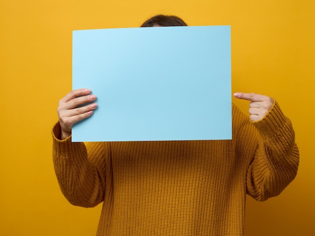 Mulher de suéter laranja segura uma folha de papel em branco sobre fundo amarelo Lugar para uma informação de anúncio de inscrição