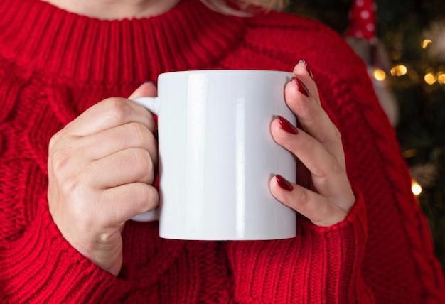 Mulher de suéter vermelho com xícara branca nas mãos contra a árvore de Natal com luzes mock up