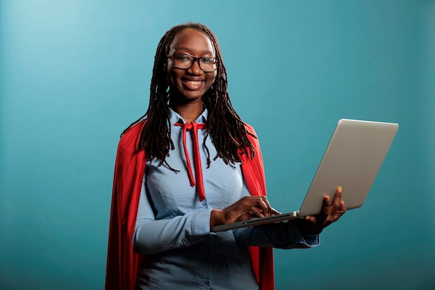 Mulher de super-herói alegre feliz vestindo manto vermelho poderoso herói em pé sobre fundo azul. Retrato de defensor da justiça americano africano confiante e positivo navegando na internet no laptop moderno.