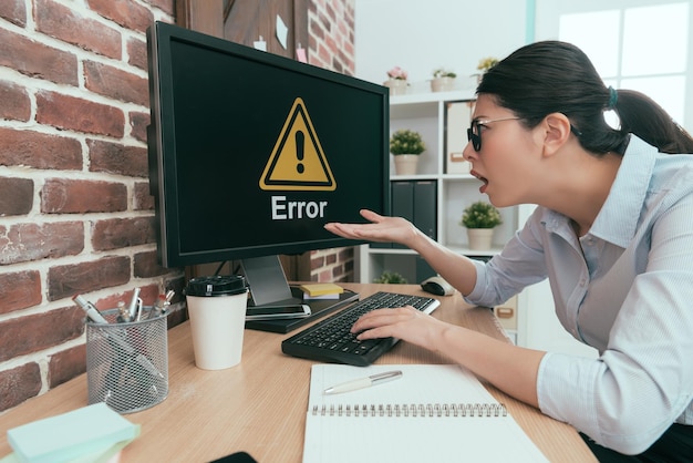 mulher de terno de tristeza sentada na mesa de trabalho usando o teclado digitando fazendo relatório, mas o computador mostrando informações de erro deixa ela se sentindo pasma.