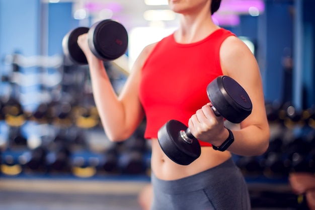 Mulher de top vermelho com cabelo loiro curto, treinando bíceps com halteres no ginásio. Conceito de pessoas, fitness e estilo de vida