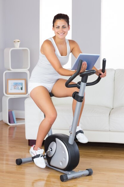 Mulher de treinamento feliz usando uma bicicleta de exercício enquanto segura um comprimido