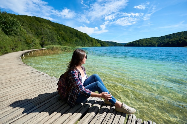 Mulher de turista sentado na ponte de madeira, apreciando a bela vista, panorama colorido da primavera de iluminado pela floresta verde sol e lago azul sob o céu claro