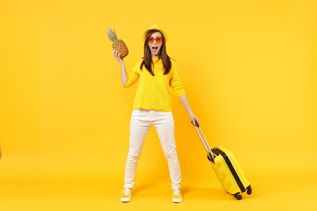 Mulher de turista viajante excitada em roupas de verão que seguram frutas frescas de abacaxi maduras isoladas em fundo laranja amarelo. Passageiro viajando para o exterior em escapadelas de fim de semana. Conceito de viagem de voo aéreo.