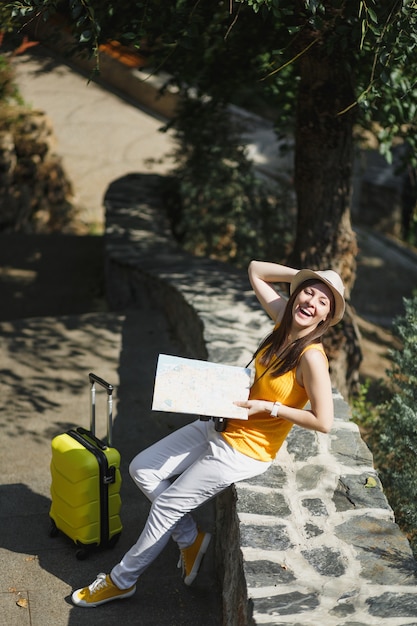 Mulher de turista viajante feliz rindo com chapéu de roupas amarelas com mapa da cidade mala sentado na pedra no parque da cidade ao ar livre. garota viajando para o exterior para viajar no fim de semana. estilo de vida da viagem de turismo.