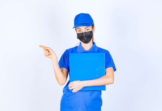 Mulher de uniforme azul e máscara preta, segurando uma pasta azul e apontando para alguém à parte.