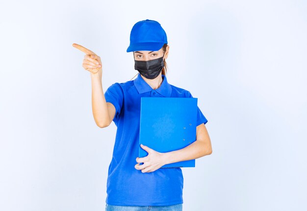 Mulher de uniforme azul e máscara preta, segurando uma pasta azul e apontando para alguém à parte.