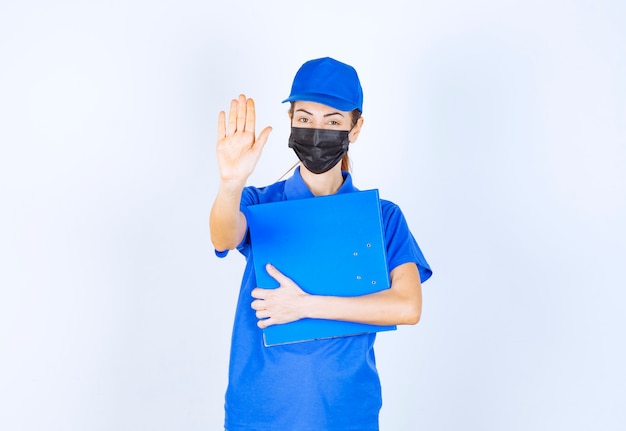 Mulher de uniforme azul e máscara preta, segurando uma pasta azul e parando alguma coisa.