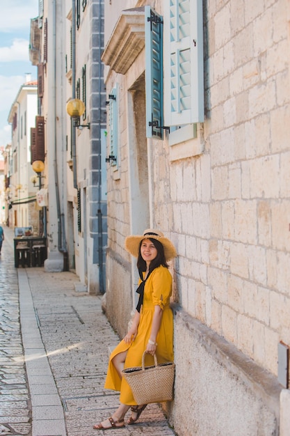 Mulher de vestido amarelo de verão com chapéu de pé perto do edifício de pedra na cidade resort de verão