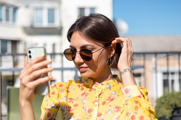 Foto mulher de vestido amarelo no terraço de um café de verão com telefone celular em dia de sol