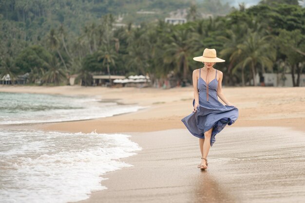 Mulher de vestido azul e chapéu de palha anda na praia