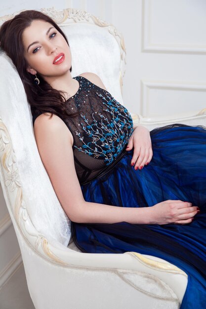 Mulher de vestido azul posando enquanto está sentado no sofá vintage e olhando para a câmera com sorriso