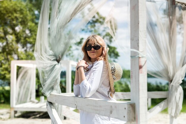 Mulher de vestido branco aproveite o dia quente de verão perto da praia de areia no gazebo de madeira branco. férias ou relaxe