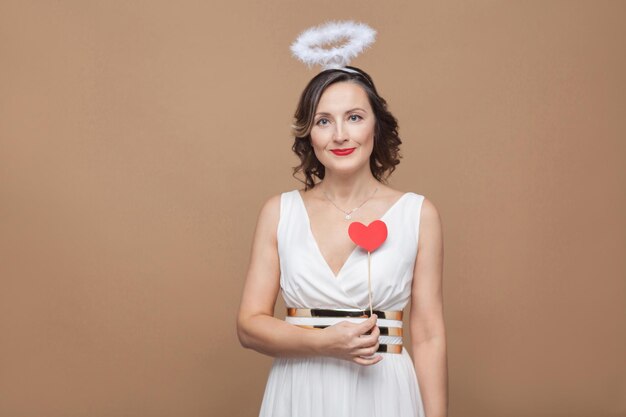 Mulher de vestido branco com nimbo na cabeça segurando um adesivo de coração vermelho e olhando para a câmera com um sorriso