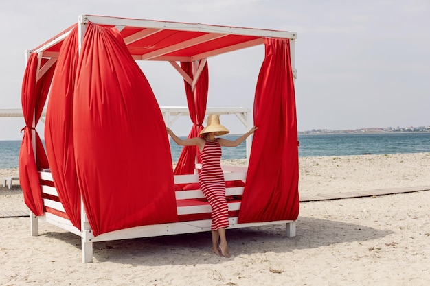 Foto mulher de vestido listrado vermelho está ao lado de uma cama de sol coberta em uma praia de areia