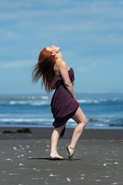 Mulher de vestido longo de pé na praia de areia com a cabeça olhando para cima e os olhos fechados tomando banho de sol