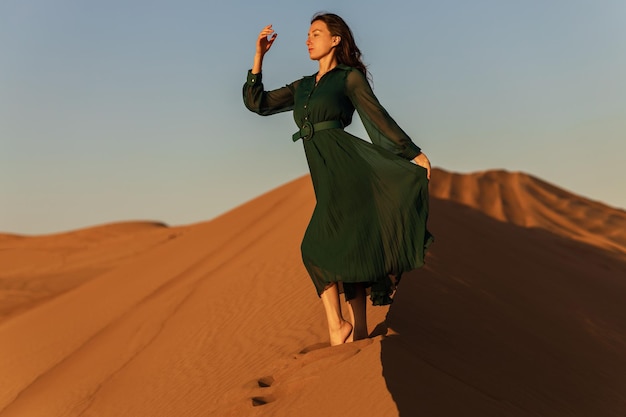 Mulher de vestido longo verde fica em fundo de dunas de areia do deserto amarelo laranja pôr do sol