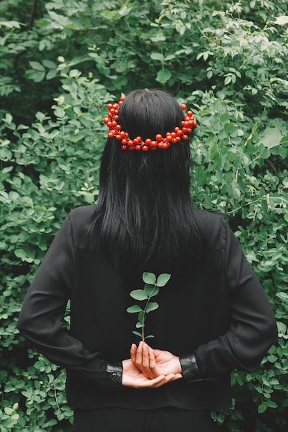 Mulher de vestido preto e enfeites vermelhos segurando um galho na mão contra o fundo da floresta