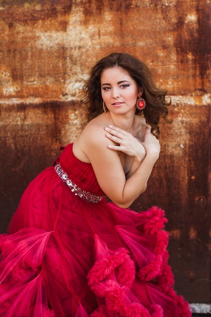 Mulher de vestido vermelho nublado está posando sobre parede de ferrugem. Retrato da moda sem retoque, apenas maquiagem