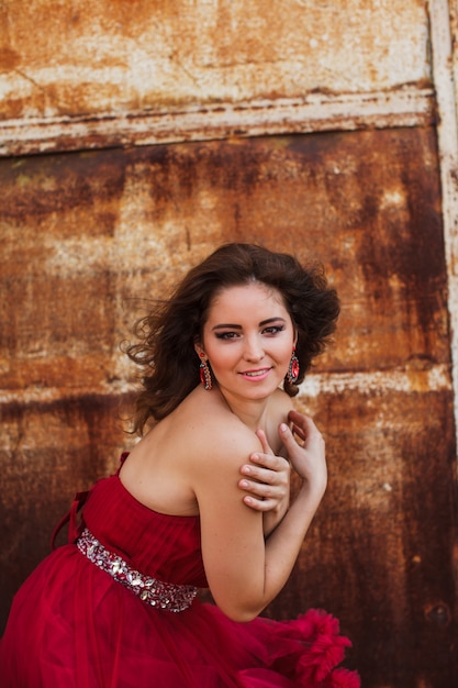 Mulher de vestido vermelho nublado está posando sobre parede de ferrugem. Retrato da moda