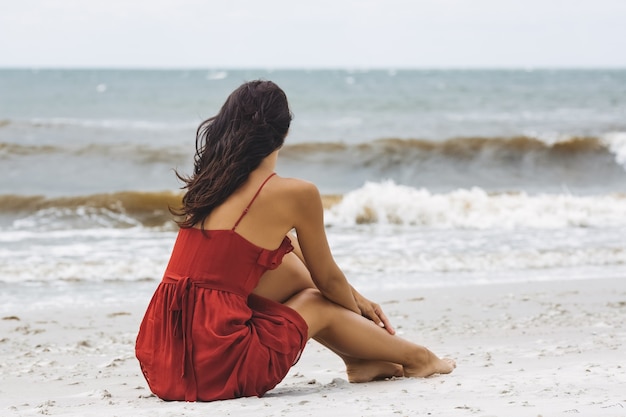 Mulher de vestido vermelho sentada na areia em um clima frio e ventoso