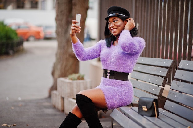 Mulher de vestido violeta e boné posou ao ar livre, sentado no banco e fazendo selfie no telefone.