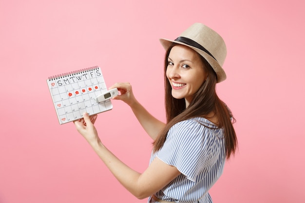 Mulher de vista traseira traseira em vestido segurar termômetro na mão, calendário de períodos femininos, verificando dias de menstruação isolados no fundo rosa. Saúde médica, conceito ginecológico de ovulação. Copie o espaço