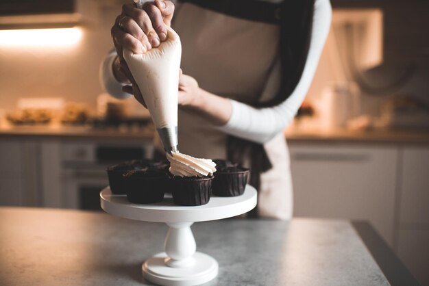 Mulher decora muffins de chocolate com queijo de creme batido na mesa da cozinha em close-up trabalhando em casa