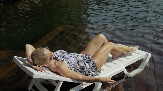 Mulher deitada em uma espreguiçadeira em óculos de sol e um xale de seda boho Menina descansa em um cais subaquático de madeira de inundação O pavimento está coberto de água no lago