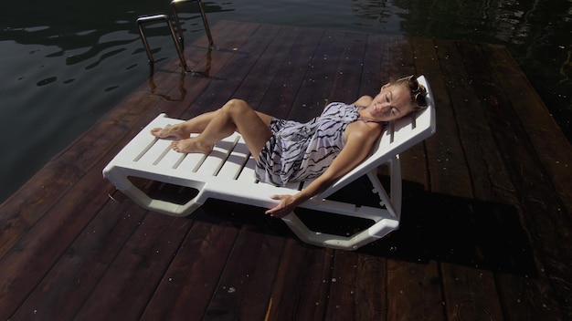 Mulher deitada em uma espreguiçadeira em óculos de sol e um xale de seda boho Menina descansa em um píer subaquático de madeira de inundação O pavimento está coberto de água no lago