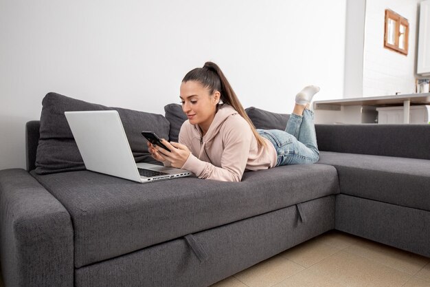 Foto mulher deitada no sofá olhando para o smartphone na frente do laptop