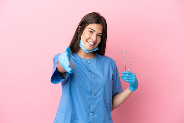 Mulher dentista segurando ferramentas isoladas em um fundo rosa apertando as mãos para fechar um bom negócio