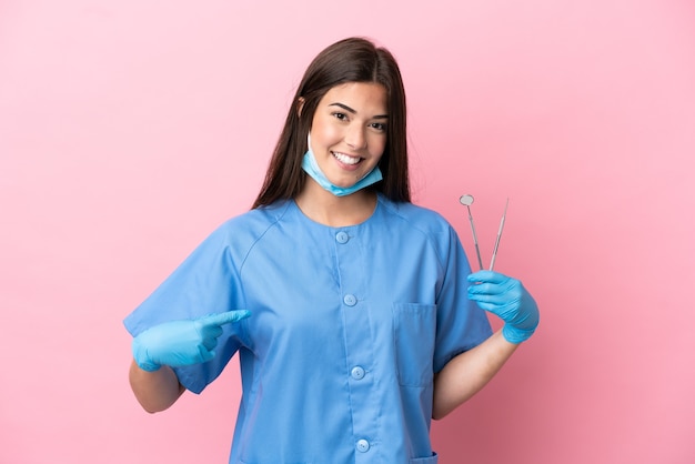 Mulher dentista segurando ferramentas isoladas em um fundo rosa com expressão facial surpresa