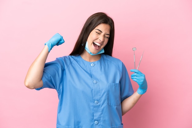 Mulher dentista segurando ferramentas isoladas em um fundo rosa, comemorando uma vitória