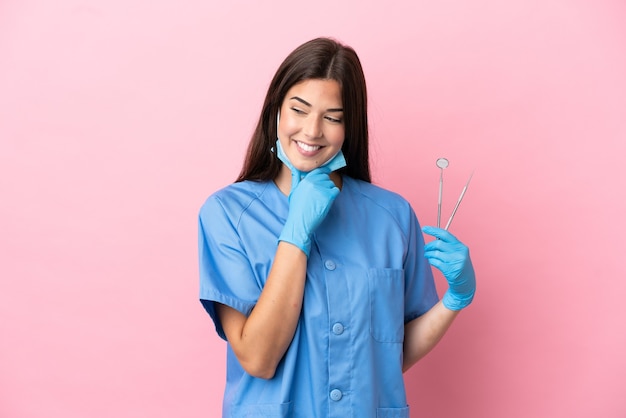 Mulher dentista segurando ferramentas isoladas em um fundo rosa, olhando para o lado e sorrindo