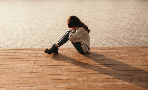 mulher deprimida sentada sozinha em um píer de madeira perto do lago