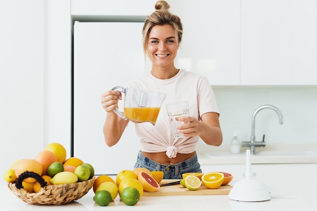 Mulher derramando suco de laranja caseiro espremido na hora no copo na cozinha branca moderna