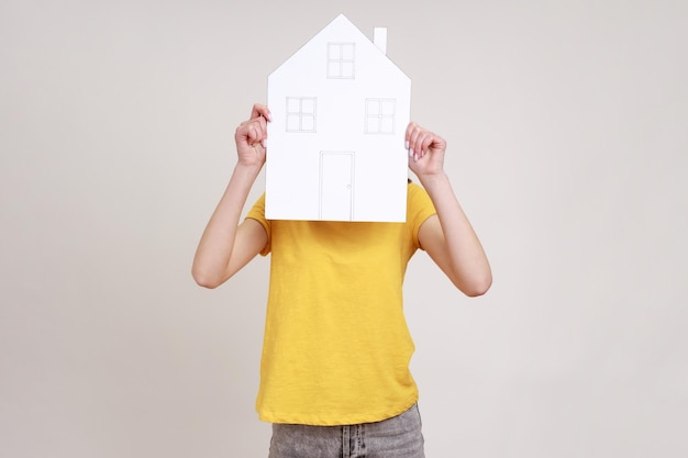 Mulher desconhecida em camiseta amarela escondendo o rosto atrás da casa de papel com janelas desenhadas e programa de habitação do governo de porta sonhando com casa nova Tiro de estúdio interior isolado em fundo cinza