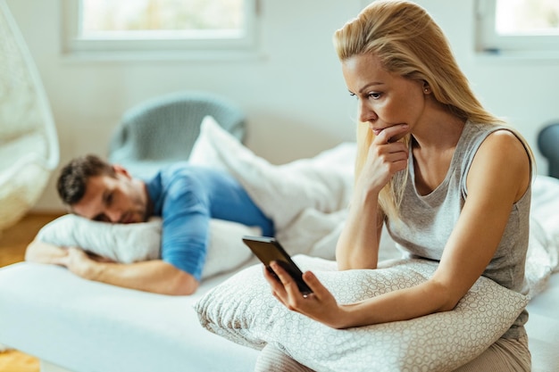 Mulher descontente lendo mensagem de texto no celular enquanto o namorado está dormindo na cama ao fundo.