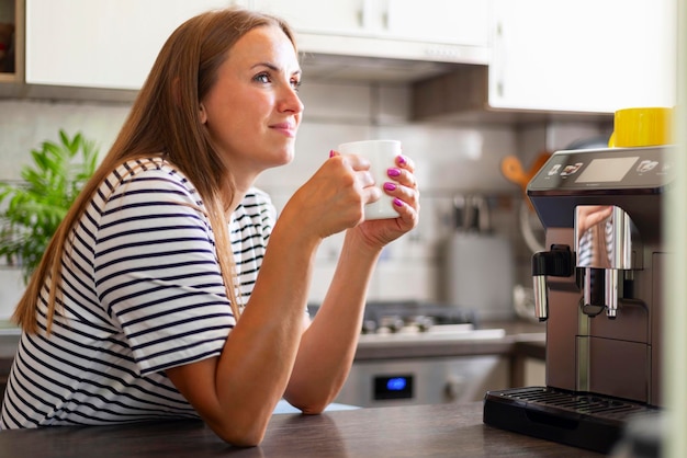 Mulher desfrutando de café recém-preparado perto de uma máquina de café moderna na cozinha